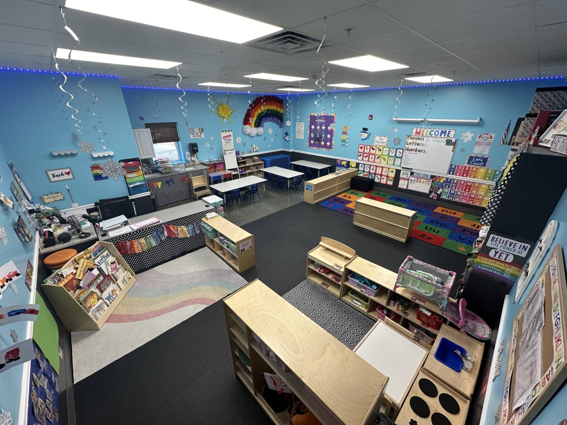 A bright and colorful preschool classroom, filled with small, child-sized furniture. The room features a variety of learning stations, including a reading corner with a small bookshelf stocked with picture books, a play area with educational toys, and an arts and crafts table covered with paper, crayons, and paints. The walls are decorated with children's artwork and educational posters showcasing the alphabet, numbers, and basic shapes. Natural light streams in from large windows, and a cozy rug lies in the center of the room for group activities. The atmosphere is cheerful and inviting, designed to stimulate learning and creativity in young children.