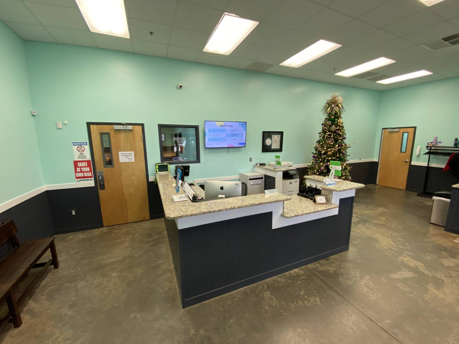 A welcoming front desk reception area in a facility, with a tranquil aqua-colored wall backdrop. The L-shaped reception counter is finished in dark blue with speckled granite countertops, housing desktop computers, telephones, and organizational compartments for efficient guest service. A wooden bench sits to the left, providing a waiting area alongside a prominently displayed safety notice. To the right, a decorated Christmas tree brings seasonal warmth to the space. Stained concrete flooring underpins the area, offering a modern and clean look. Overhead, bright fluorescent lights ensure a well-lit environment. Dual wooden doors with glass panels flank the desk, suggesting entry points for staff and visitors, while an informational screen above the desk keeps guests informed. The setup exudes a professional yet inviting atmosphere for all who enter.