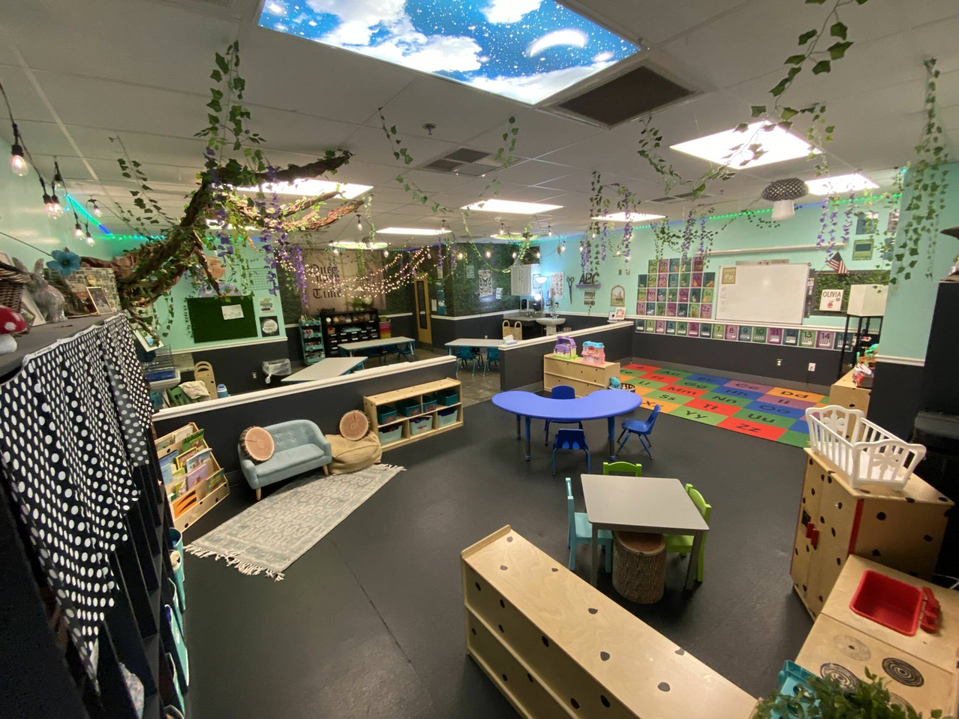 An inviting and vibrant preschool classroom with an imaginative play theme. The ceiling is adorned with strings of lights and decorative greenery, simulating an enchanted forest. The floor is covered with a multicolored alphabet carpet and various play areas. To the left, a cozy reading nook with a small blue sofa, bookshelf, and polka dot curtain invites quiet time. In the center, a blue circular table and a green square table provide spaces for group activities. The room's periphery features learning stations, including a whiteboard, a bulletin board with children's names, and a kitchen playset. An interactive corner with a crib and wooden play structures encourages imaginative play. The space is well-lit, with both fluorescent lighting and a starry sky painted ceiling, creating a magical learning atmosphere.