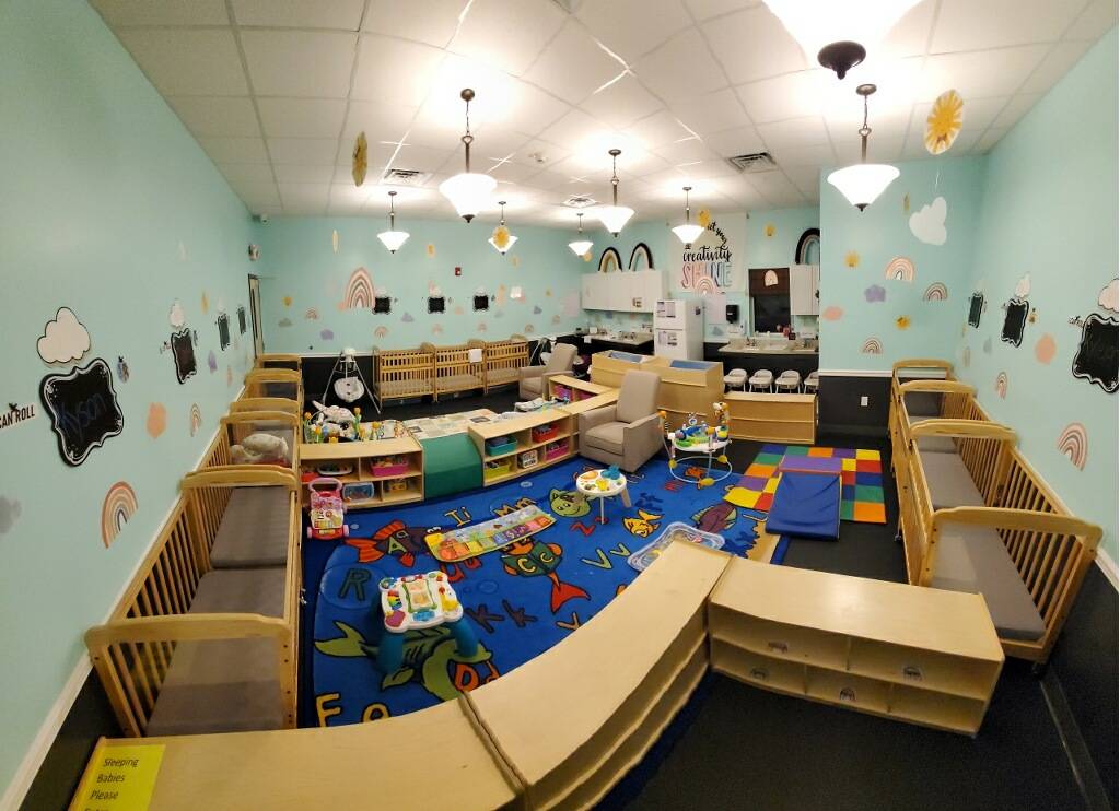 A warm and soothing infant room designed for comfort and safety. The room is equipped with multiple cribs, each with soft bedding and a mobile hanging above. The color palette is gentle, with pastel walls and soft lighting to create a calm atmosphere. In one corner, there's a cozy rocking chair for feeding or comforting the infants. Along another wall, there are shelves stocked with age-appropriate toys and sensory materials, like soft blocks, rattles, and plush animals. A changing station is set up for convenience, complete with all necessary supplies. The floor is covered with a clean, soft mat for supervised tummy time and crawling. The room is meticulously maintained to ensure a hygienic and nurturing environment for the youngest children.